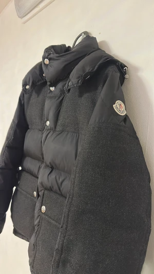 Svart Moncler pufferjacka med ull - Säljer en svart pufferjacka från Moncler med coola ullpaneler och blanka knappar. Jackan har hög krage, långa ärmar och två stora fickor framtill. Moncler-loggan sitter på ärmen och materialmixen ger en lyxig vibe. Perfekt för dig som vill ha något unikt och varmt.