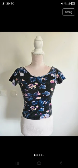 Svart blommig croppad topp - Säljer en svart croppad topp med blommigt mönster i blått, lila och rosa toner. Toppen har korta ärmar och rundad halsringning både fram och bak. Materialet känns mjukt och stretchigt, perfekt för dig som gillar färg och mönster.