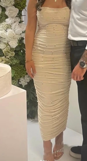 Champagnefärgad pärlklänning midi  - Säljer en elegant champagnefärgad långklänning med smala axelband. Klänningen är figurnära med draperade detaljer och dekorerad med små pärlor över hela tyget. Perfekt för dig som vill sticka ut med en lyxig och feminin look. Klänningen är bekväm och endast använd några timmar, fortfarande som ny 