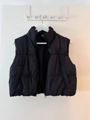 Svart pufferväst med dragkedja - Svart pufferväst med hög krage och praktiska fickor på sidorna. Västen har en rejäl dragkedja framtill och är tillverkad i ett syntetmaterial som ger en puffig look. Perfekt att slänga över hoodien för extra värme och stil.