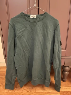 Grön sweatshirt från Stone Island - Säljer en grön långärmad sweatshirt från Stone Island med klassisk rund hals och ikonisk patch på ärmen. Tröjan är i mjuk bomull och har ribbade muddar vid ärmslut och nederkant. Perfekt för dig som gillar streetwear och snygga detaljer.