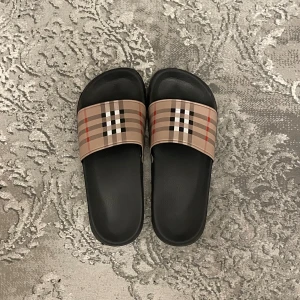 Burberry tofflor - Fina Burberry sandaler med bred rem i beige och klassiskt rutmönster kommer fint packade. Snygg och enkel design med platt sula och öppen tå, pris kan diskuteras vid snabb affär.