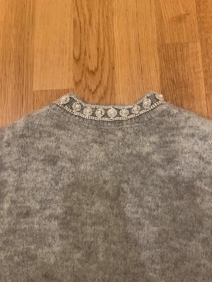 Grå kofta med pärlor från Zara - Superfin grå kofta från Zara med glittrande pärldetaljer och strass vid halsen. Koftan är stickad och har knappar framtill samt ribbade muddar vid ärmar och nederkant. Perfekt för dig som gillar att sticka ut med snygga detaljer. Vissa diamanter är borta men inget man tänker på.