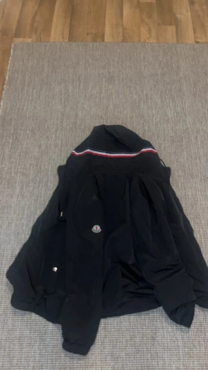 Svart vindjacka från Moncler - Svart vindjacka från Moncler med huva och klassisk logga på bröstet. Jackan har en diskret röd, vit och blå rand över ryggen och är tillverkad i lätt polyester. Perfekt för dig som gillar stilrena och sportiga plagg med premiumkänsla.