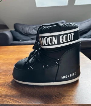 Svarta Moon Boots med snörning - Moon Boot i svart med vit logga och snörning framtill. Skon har en rund tå och platt sula, perfekt för kalla vinterdagar. Materialet är syntet och designen är klassisk med högt skaft och tydlig Moon Boot-märkning.