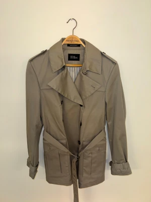 Beige trenchcoat från Oscar Jacobson - Snygg beige trenchcoat från Oscar Jacobson med klassisk krage, axelklaffar och bälte i midjan. Jackan har långa ärmar, knappar framtill och praktiska fickor. Insidan är fodrad med ett diskret randigt mönster. Perfekt för dig som gillar stilrena och tidlösa plagg.
