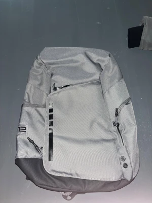 Nike Elite grå ryggsäck - Snygg grå Nike Elite ryggsäck med svarta detaljer och flera dragkedjefack. Väskan har justerbara axelremmar med transparenta paneler och en tydlig Elite-logga på framsidan. Tillverkad i slitstark polyester, perfekt för att bära allt du behöver till skolan eller träningen.