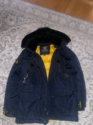 Mörkblå dunjacka Geographical Norway M - Säljer en mörkblå vinterjacka från Geographical Norway i storlek M. Jackan har gul quiltad insida, flera fickor med dragkedja och snygga detaljer i svart. Stor avtagbar huva med fuskpäls och justerbara spännen. Varmaste jackan jag har haft. Insidan av jackan är lite missfärgad därav nedsänkt pris.