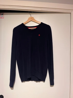 Mörkblå merinoullströja från Morris - Snygg mörkblå tröja från Morris i 100% merinoull. V-ringad modell med lång ärm och diskret röd broderad logga på bröstet. Perfekt för dig som gillar klassisk stil med en modern touch.