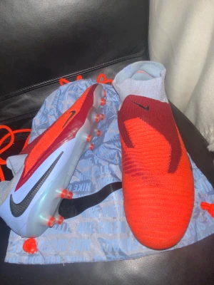 Nike röda fotbollsskor med strumpkrage - Säljer ett par grymma Nike fotbollsskor i rött och vinrött med ljusgrå strumpkrage. Skorna har en platt sula med dobbar för bra grepp på planen och är tillverkade i syntetmaterial med stickad ovandel. Snygg design med svart Nike-logga på sidan och ovansidan.