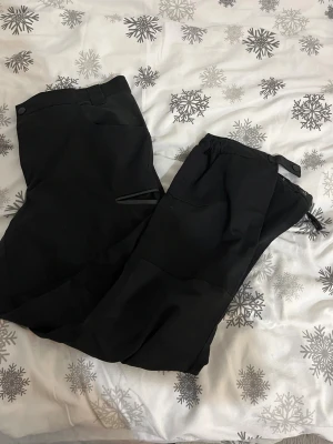 Svarta täckbyxor med snörning - Svarta täckbyxor med praktiska fickor och snörning vid benslut. Perfekta för kalla dagar och utomhusaktiviteter. Materialet är slitstarkt och håller dig varm. Snygg och enkel design som passar till många jackor.