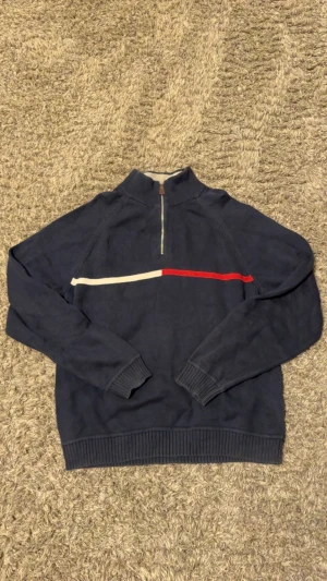 Marinblå half zip tröja Tommy Hilfiger - Snygg marinblå stickad tröja från Tommy Hilfiger i storlek M. Tröjan har en half zip-dragkedja och en vit samt röd rand över bröstet. Perfekt för dig som gillar klassisk stil med en sportig touch.