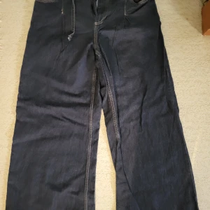 Vida mörkblå jeans med hög midja - Snygga mörkblå jeans med vida ben och hög midja. Femficksmodell med sydda veck framtill och gylf med dragkedja och knapp. Klassisk jeanslook med tillhörande smalt bälte och metallspänne. Perfekt för dig som gillar loose fit och bredare siluett.