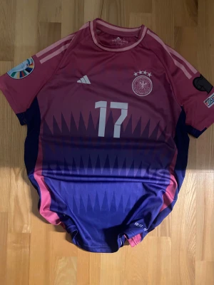 Tysklands Wirtz matchtröja rosa/lila - Säljer en officiell Tyskland fotbollströja från Adidas med Wirtz och nummer 17. Tröjan har korta ärmar, rosa upptill och mörkblå nedtill med taggigt mönster. Broderad landslagslogga, tre stjärnor och turneringspatch på ärmarna. Tillverkad i lätt funktionsmaterial.