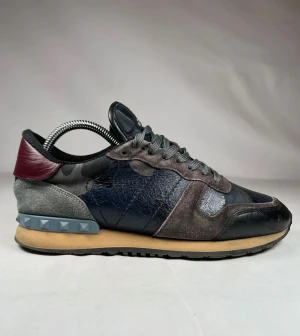 Valentino Rockrunner sneakers grå/svart - Snygga Valentino Rockrunner sneakers med coolt kamouflagemönster i grått, blått och svart. Skorna har detaljer i mocka och skinn, grå snörning och ikoniska nitar på hälen. Mörkröd detalj bak och beige sula ger extra edge. Perfekt för dig som gillar exklusiv streetstyle.