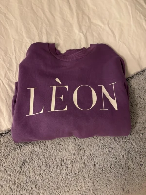 Lila sweatshirt LÈON från Gina Tricot - Cool lila sweatshirt från Gina Tricot i storlek XXS med stor vit LÈON-print framtill. Tröjan har rund hals, ribbade muddar vid ärmslut och nederkant samt lång ärm. Perfekt för dig som gillar statement-plagg och snygga färger.