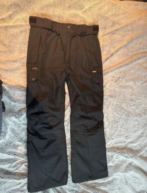 Svarta täckbyxor från Dope - Svarta täckbyxor från Dope med flera fickor, snörning i midjan och kardborre för extra justering. Byxorna har bred passform och är tillverkade i slitstarkt syntetmaterial, perfekta för kalla dagar och utomhusaktiviteter. Snygg och funktionell design med dragkedja och tryckknappar.