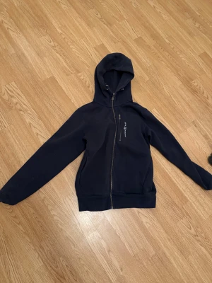 Mörkblå Sail Racing hoodie med dragkedja - Snygg mörkblå hoodie från Sail Racing med två dragkedjor framtill, varav en till bröstficka. Tröjan har huva med snörning och långa ärmar. Materialet är mjuk bomull och polyester, perfekt för chill dagar. Loggan är broderad på bröstet.