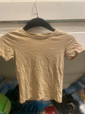 Beige kortarmad topp från 157 - En basic beige topp från 157 i 100% bomull. Modellen är enkel med rund halsringning och korta armar, perfekt att matcha med jeans eller kjol. Färgen är neutral och passar till det mesta. Snygg och stilren för en clean look. Barn till tjejer, storlek 120