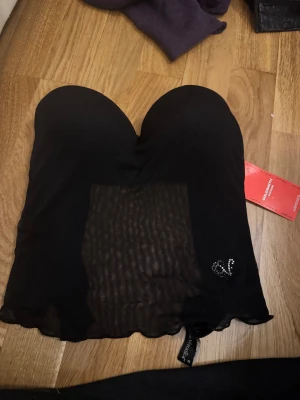 Svart mesh korsett från Verde Veronica - Säljer en svart korsett-topp från Verde Veronica med meshdetaljer och vadderade kupor. Toppen är axelbandslös och har en liten strasslogga framtill.  Så snygg på och vid bysten! Passar s/m ! Köpt i london