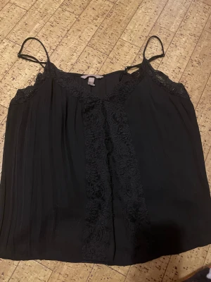 Svart spetsdetaljerad linnetopp H&M - Svart linnetopp från H&M med tunna axelband och snygga spetsdetaljer längs urringningen och framtill. Toppen har en lätt plisserad struktur och är perfekt för dig som gillar en stilren men ändå romantisk look.