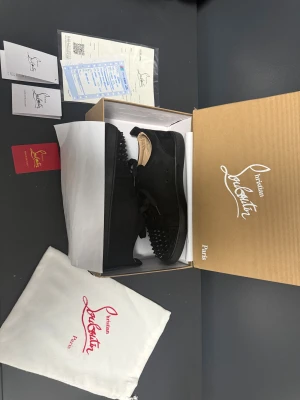Christian Louboutin skor - Svarta sneakers från Christian Louboutin i mocka med coola svarta nitar på tån och klassisk röd sula. Skorna har rund tå, snörning och platt sula. Insidan är beige och de kommer med originalkartong och dustbag. Perfekt för dig som vill sticka ut med lyxig streetstyle. Pris kan diskuteras eftersom jag använt dem en gång.
