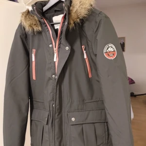 Svart parkajacka med fuskpäls splitter ny Medium - Svart parkajacka med avtagbar huva och fuskpäls. Jackan har orange dragkedjor, två bröstfickor, stora fickor framtill och patch på ärmen. Vadderad och perfekt för kalla dagar. Snygg och funktionell design med hög krage. Splitter ny Medium normal passform.