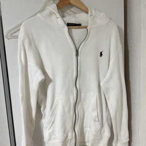 Vit hoodie från Polo Ralph Lauren - Vit hoodie från Polo Ralph Lauren med dragkedja och huva. Tröjan har broderad svart logga på bröstet, två fickor fram och är tillverkad i mjuk bomull. Storlek S men passar M