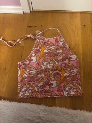 Färgglad halterneck topp - Säljer en snygg halterneck topp med knyt i nacken. Toppen har ett paisleyinspirerat mönster i rosa, orange och vitt. Perfekt för dig som gillar färg och mönster! Materialet känns lätt och skönt mot huden.