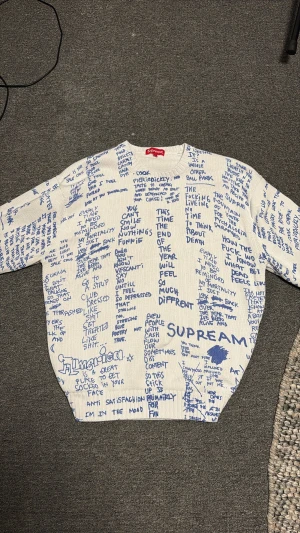 Vit Supreme stickad tröja med blå text - Supreme stickad tröja i storlek L, vit med blå handskrivna texter över hela plagget. Crewneck-modell med ribbade muddar och nederkant. Tillverkad i 100% bomull, riktigt unik design som sticker ut och passar dig som gillar streetwear. Självklart äkta