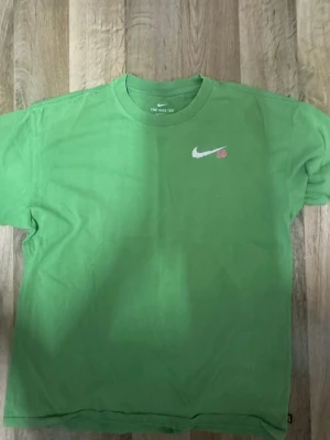 Grön Nike SB t-shirt med tryck - Grön Nike SB t-shirt i bomull med klassisk rund hals och korta ärmar. Framsidan har en liten vit Nike SB-logga, medan baksidan har ett stort tryck med en gul drake, moln och rosa Nike SB-text. Perfekt för dig som gillar streetwear och coola prints.