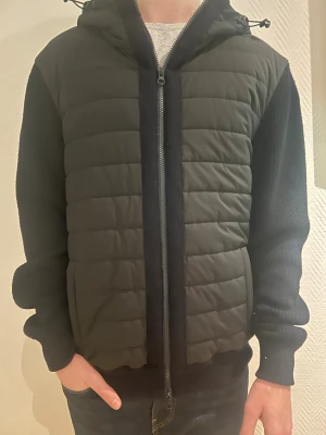 Svart Pufferjacka/Cardigan från Gran Sasso L/M - Hej 👋 säljer en snygg svart pufferjacka från Gran Sasso med stickade ärmar och quiltad framsida. Jackan har huva med snörning och dragkedja framtill. Tillverkad i Rain Wool, vilket gör den extra tålig mot väta. Perfekt för dig som vill ha en modern och bekväm look. Vid frågor och funderignar kan du kontakta mig