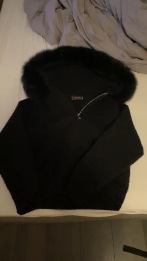 Svart päls jacka från Alessa - Svart jacka från Alessa med fluffig huva och pälsdetaljer runt kanten. Jackan har lång ärm och en snygg dragkedja framtill. Perfekt för dig som gillar en mysig och trendig look under höst och vinter. 