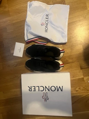 Svarta sneakers från Moncler - Säljer ett par svarta chunky sneakers från Moncler med vit och svart sula samt Monclers signaturrand i rött, vitt och blått på hälen. Skorna har snörning och är tillverkade i en mix av syntet och skinn. Kommer med originalkartong och dustbag. Dem är lite stora i Storleken så rekommendation är 43 