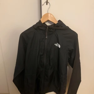 Svart vindjacka från The North Face XL (barn) - Svart vindjacka från The North Face i storlek XL (barnstorlek). Jackan har huva, dragkedja framtill och logga på både bröstet och ryggen. Tillverkad i lätt polyester, perfekt för blåsiga dagar. Snygg och enkel design med långa ärmar och praktiska fickor.