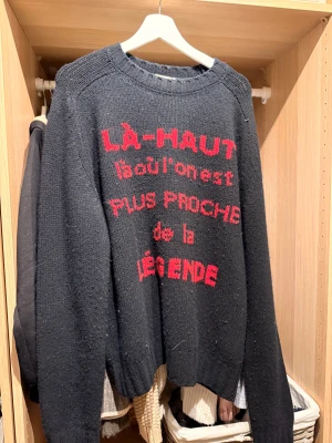 Svart stickad Moncler tröja med röd text - Svart stickad tröja från Moncler med röd text på framsidan: 'LÀ-HAUT làoù l'on est PLUS PROCHE de la LÉGENDE'. Tröjan har Moncler-logga på ärmen, rund hals och långa ärmar. Perfekt för dig som gillar statement-plagg och exklusiva märken.