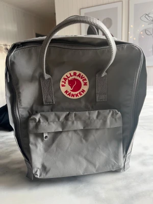 Grå Fjällräven Kånken ryggsäck - Säljer en ny oanvänd klassisk grå Fjällräven Kånken ryggsäck med det ikoniska röda logotypmärket framtill. Väskan har ett stort huvudfack, en praktisk frontficka med dragkedja och två handtag upptill. Tillverkad i slitstark syntet och har justerbara axelremmar.