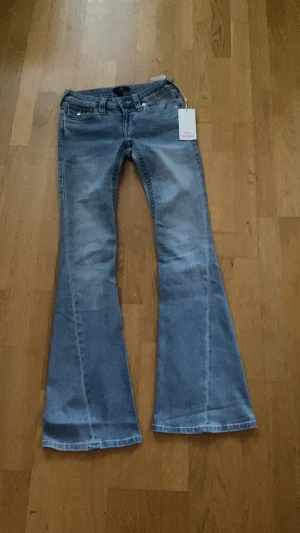 True Religion Joey Flare Jeans blå - Blå jeans från True Religion, modell Joey Low Rise Flare. Snygg låg midja och utställda ben med klassisk femficksdesign och detaljerade bakfickor med knappar. Materialet är jeans och färgen är ljusblå med tvättade partier för cool effekt.