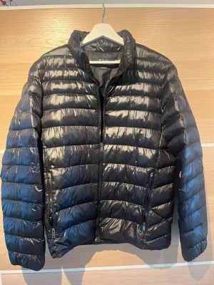 Svart pufferjacka från Polo Ralph Lauren - Snygg svart pufferjacka från Polo Ralph Lauren i storlek M. Jackan har horisontella quiltade paneler, hög krage och två sidofickor med dragkedja. Materialet är glansigt och känns lätt, perfekt för kyliga dagar. Klassisk och stilren design med diskret logga på insidan. Ny pris tror jag var 3800 mitt pris 999