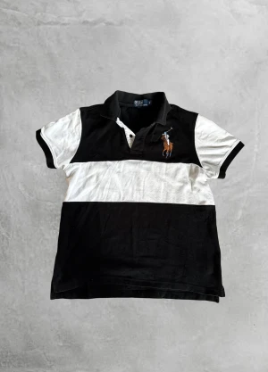 Svartvit piké från Polo Ralph Lauren - Klassisk pikétröja från Polo Ralph Lauren i svart och vitt med breda blockränder. Tröjan har broderad polospelare på bröstet, krage och knappar framtill. Tillverkad i mjuk bomull och har korta ärmar. Perfekt för en sportig och stilren look.