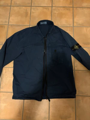 Marinblå overshirt från Stone Island - Säljer en marinblå overshirt från Stone Island med klassisk patch på ärmen, bröstficka med knapp och kraftig dragkedja framtill. Riktigt fet overshirt som jag tyvärr inte använder längre.