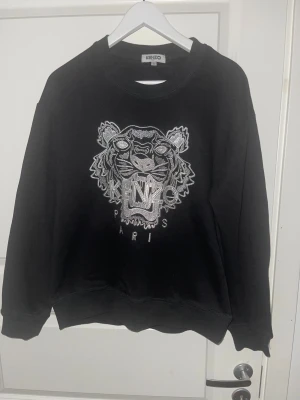 Svart sweatshirt från Kenzo med tiger - Svart sweatshirt från Kenzo med ikonisk broderad tiger i silvers på bröstet och texten 'Kenzo Paris'.
