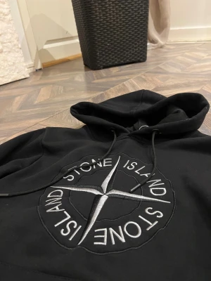 Svart hoodie från Stone Island - Svart hoodie från Stone Island med stort broderat logomärke i vitt och grått på bröstet samt klassisk patch med gul och grön kompass på ärmen. Tröjan har huva med snörning och är tillverkad i mjuk bomull. Perfekt för dig som gillar streetwear och snygga detaljer.