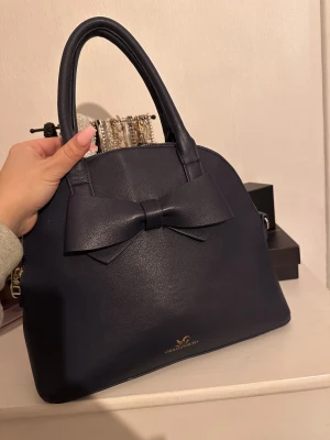 Mörkblå handväska med rosett Holly & Whyte - Säljer min  mörkblå handväska från Holly & Whyte i skinnimitation med en stor rosett framtill. Väskan har rundad form, guldfärgade detaljer och dragkedja upptill. 