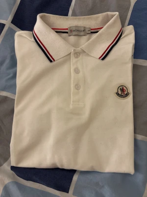 Vit Moncle pikétröja - Snygg vit pikétröja från Moncle med klassisk krage och knäppning framtill. Kragen och ärmsluten har stilrena rödblå ränder. Tröjan har Moncler-logga broderad på bröstet och är gjord i mjuk bomull. Perfekt för dig som gillar clean och sportig stil. Strl m men passar även för L