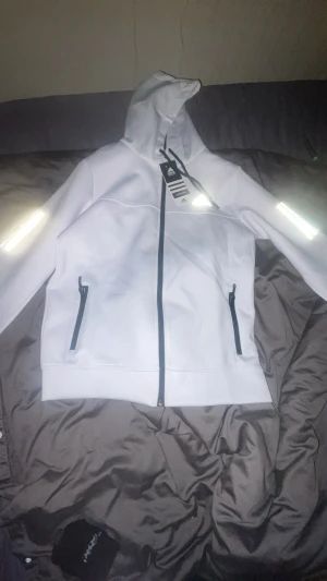 Vit vindjacka med huva från Adidas XS - Snygg vit vindjacka från Adidas i storlek XS. Jackan har huva med snörning, svarta dragkedjor på fickorna och framtill, samt reflekterande detaljer på ärmarna. Materialet är syntet och jackan har en clean, sportig look som passar perfekt till streetwear.