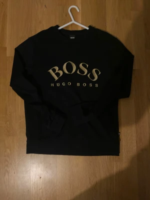 Svart sweatshirt från Hugo Boss - Svart sweatshirt från Hugo Boss med guldig BOSS-logga framtill. Klassisk rund halsringning och långa ärmar. Tröjan är i mjuk bomullsmix och har ribbade muddar vid ärmslut och nederkant. Perfekt för en clean och stilren look. Storlek S-M