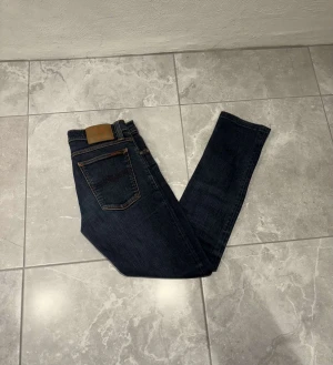 Mörkblå jeans från Nudie Jeans - Snygga mörkblå jeans från Nudie Jeans med brunt läderpatch bak i midjan. Jeansen har en rak och slim passform som sitter schysst på benen. Perfekta för dig som gillar stiliga jeans. Storleken är W28 och L32