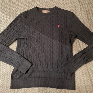 Svart kabelstickad tröja Morris S - Blå kabelstickad tröja från Morris i 100% merinoull. Klassisk rund hals och långärmad modell med ribbade muddar. Diskret röd broderad logga på bröstet ger en snygg detalj. Perfekt för dig som gillar stilrena och bekväma plagg.