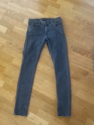 Grå skinny jeans från Lee Malone W31 L32 - Snygga grå jeans från Lee, modell Malone, med smal passform . Jeansen är tillverkade i mjukt denimtyg och har en stilren look med svarta Lee-detaljer på knapp och etikett. Perfekt för dig som gillar en modern och slimmad siluett. Jeansen är i bra skick.